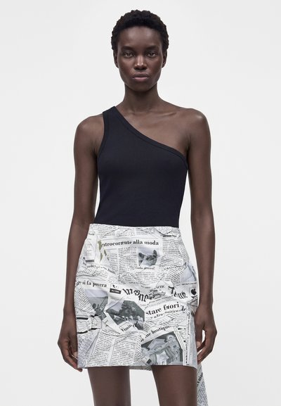 Γυναίκα που φοράει μαύρο τοπ με έναν ώμο και λευκή φούστα με τύπωμα εφημερίδας, στέκεται μπροστά σε ένα απλό φωτεινό φόντο.