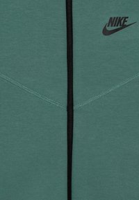 Zelená látka s černým centrálním zipem a černým logem Nike v pravém horním rohu, zdobená šikmými švy.