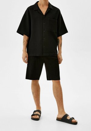 Black short-sleeve shirt with a pocket, featuring a textured fabric and a relaxed fit, paired with knee-length black shorts and black sandals.
