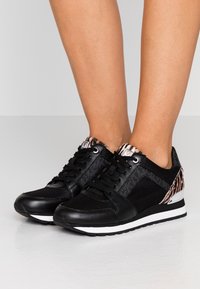 Baskets athlétiques noires en mesh et cuir, avec un talon arrière à motif et une semelle blanche avec un accent argenté.