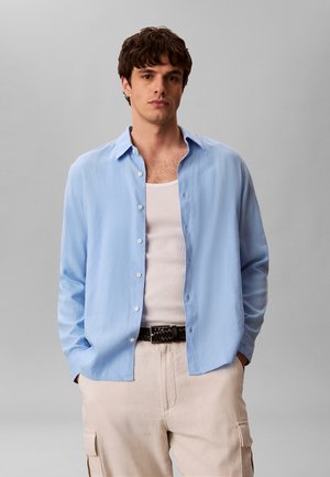 Junger Mann mit hellblauem aufgeknöpftem Hemd über einem weißen Tanktop, beige Cargohosen und einem schwarzen geflochtenen Gürtel, die Hände in den Taschen.