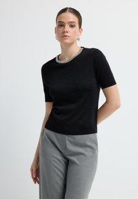 Pull noir à manches courtes avec un accent métallique au niveau du col, fabriqué en tissu texturé. Assorti avec un pantalon à rayures légères gris clair.