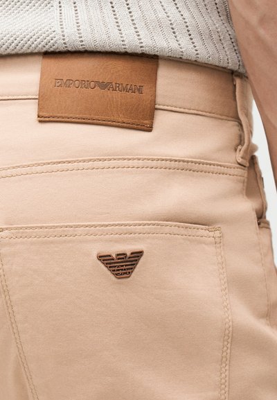 Κοντινό πλάνο μπεζ παντελονιού με καφέ δερμάτινη ετικέτα Emporio Armani στη μέση και μεταλλικό λογότυπο αετού στην πίσω τσέπη.