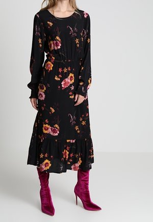 Kvinde iført en sort, langærmet midi-kjole med blomsterprint, kombineret med lyserøde velour støvler med høje hæle, stående mod en ensfarvet baggrund.