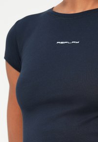 Tricou mulat de culoare bleumarin cu mâneci scurte, având logo-ul alb "REPLAY" centrat pe partea superioară a pieptului.