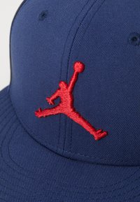 Tamsiai mėlyna kepurė su tekstūruota medžiaga; priekyje išryškėja ryškiai raudonas išsiuvinėtas Jumpman logotipas, išsiskiriantis sportiniu dizainu.