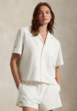 Kvinde stående med hænderne i lommerne iført hvid tekstureret kortærmet skjorte med knapper og matchende shorts mod en neutral baggrund.