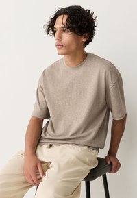 T-shirt met korte mouwen en een ronde hals in lichtbruine, zachte stof met een losse pasvorm. Gecombineerd met crèmekleurige broek, zittend op een donkere stoel.