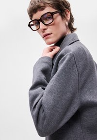 Blazer cinza texturizado com gola em bico, complementado por óculos castanhos oversized. Apresenta um corte descontraído e detalhes de costura visíveis.