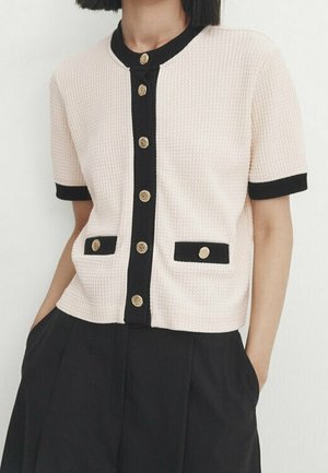 Femme portant un cardigan crème à manches courtes avec une bordure noire et des boutons dorés, associé à un pantalon noir taille haute.