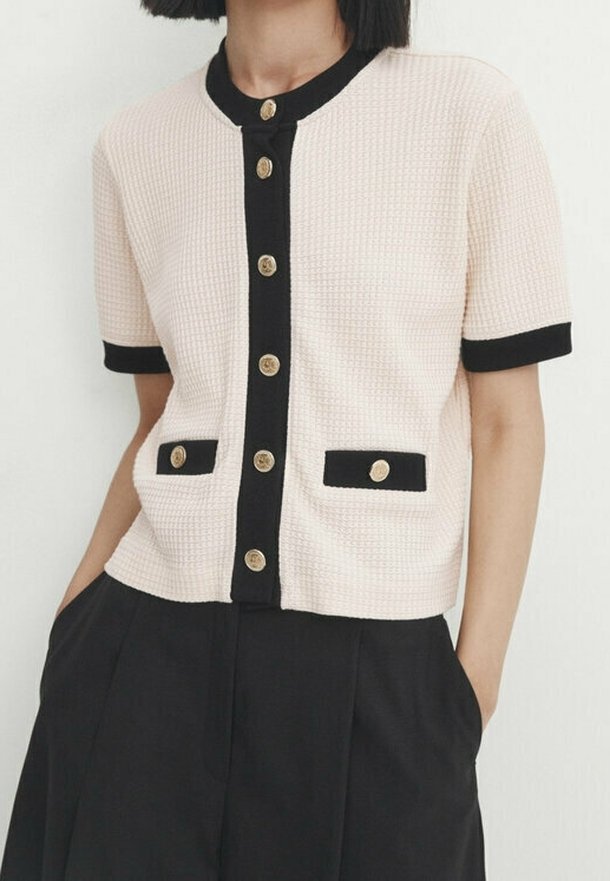 Femme portant un cardigan crème à manches courtes avec une bordure noire et des boutons dorés, associé à un pantalon noir taille haute.