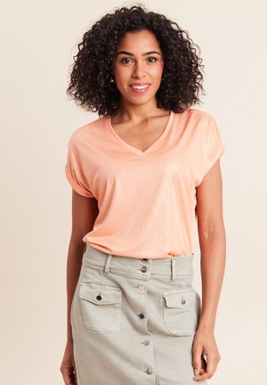 Vrouw met krullend haar, gekleed in een perzikkleurige V-hals top met korte mouwen en een hoog getailleerde beige rok met knopen aan de voorkant en klepzakken.