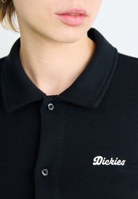 Camisa polo preta com nervuras, com frente abotoada e colarinho. Apresenta o logótipo "Dickies" bordado em branco no lado esquerdo.