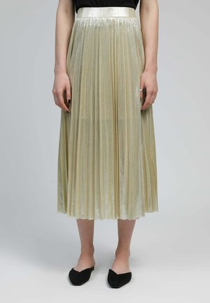 Jupe midi plissée en métal doré portée avec un haut noir et des ballerines noires pointues sur fond neutre.