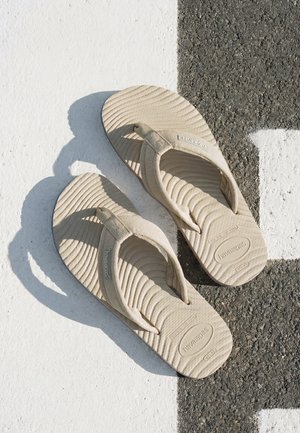 Infradito Havaianas beige con suole testurizzate posate su una superficie di pavimentazione a strisce bianche e nere alla luce del sole.