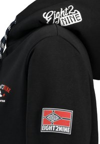 Zwarte hoodie met een gestructureerde afwerking, voorzien van een wit geborduurd logo op de capuchon en een gekleurd patch op de mouw voor branding.
