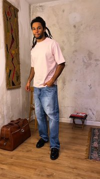 Άνδρας με ντέρεκλος, φορώντας ανοιχτό ροζ πουκάμισο και μπλε τζιν, στέκεται χαλαρά σε ένα δωμάτιο με υφασμένους τοίχους, μια ξύλινη καρέκλα, ένα δερμάτινο τσαντάκι και χαλί με σχέδια.