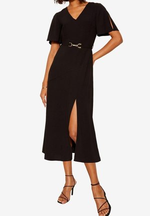 Kvinde iført en sort midi-kjole med V-udskæring, splittede ærmer, sidespalte og et dekorativt bælte, kombineret med sorte remsko med hæl.