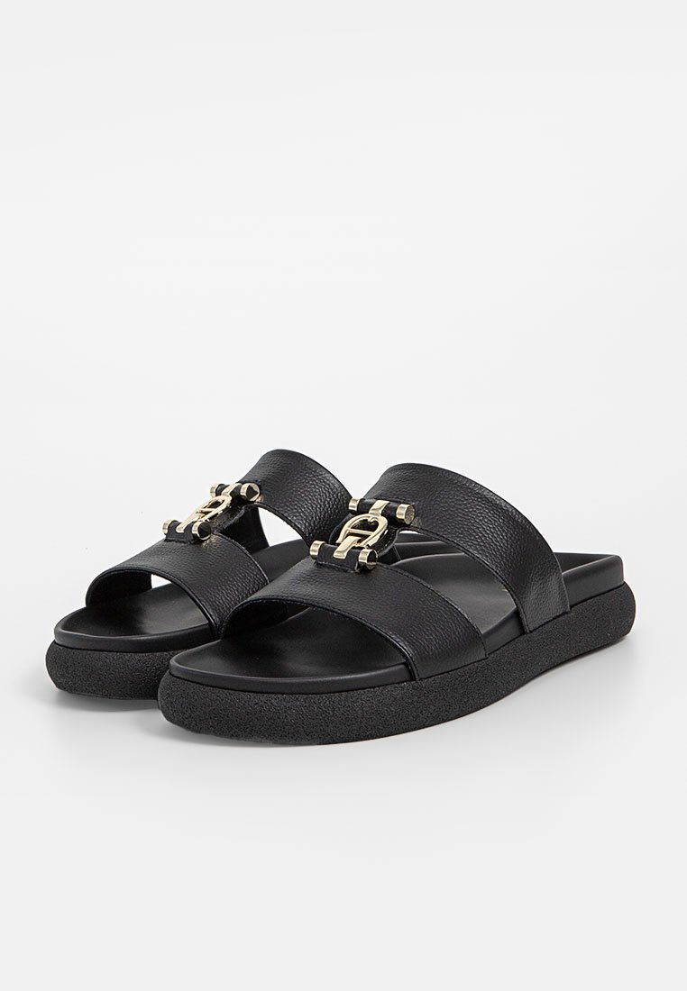 Schwarze Rutschsandalen mit zwei breiten Lederträgern, einer strukturierten Oberfläche und einem goldfarbigen Metall-Logoakzent auf der Vorderseite sowie einer klobigen Sohle.