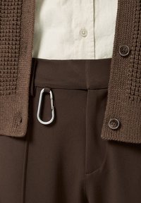 Brun stickad cardigan med stora knappar, parat med bruna byxor med en sidoficka och silverfärgad karbinhake. Off-white skjorta under.