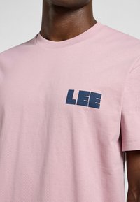 Camiseta de algodón rosa claro con cuello redondo y mangas cortas. Presenta un logo "LEE" en azul oscuro en el lado izquierdo del pecho. Textura suave.