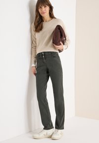 Beżowy sweter, ciemnozielone spodnie o prostych nogawkach i białe trampki. Trzyma brązową torbę, na jasnym tle.