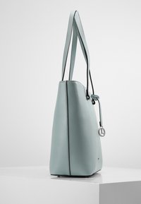 Sac fourre-tout en cuir bleu clair avec une finition texturée, deux anses, une base arrondie, des accents noirs et un détail de charme circulaire en argent.
