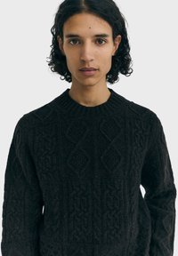 Suéter de punto marrón oscuro con un patrón texturizado que presenta formas de diamante y trenzas. Cuello redondo acanalado y mangas largas.