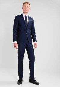 Marine dress laget av glatt stoff, med en jakke med én knapp, slim-fit bukser, og en hvit skjorte med et mørkt slips og lommefirkant.