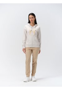 Licht beige zip-up hoodie met een gele bedrukte ontwerp, voorzakken en een trekkoord capuchon, gecombineerd met licht beige broek en witte sneakers.