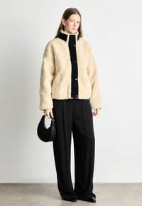 Μπεζ ψεύτικο γούνινο μπουφάν με μαύρη εμπρόσθια πάνελ, κλείσιμο με κουμπιά και ψηλό γιακά. Συνδυάζεται με φαρδιά μαύρα παντελόνια και μια μαύρη τσάντα.