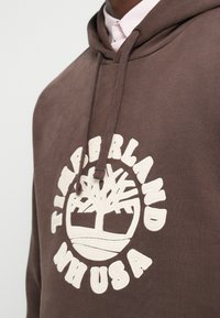 Cilvēks valkā brūnu Timberland kapuci ar balti izšūtu logotipu un auklām, kas uzlikta virs viegli rozā kreklveida krekla.