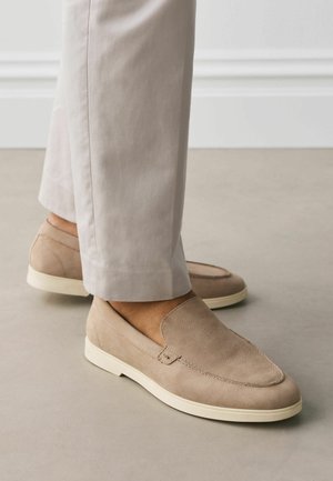 Beige Wildleder-Slipper mit weißen Gummisohlen, getragen zu hellen, gerade geschnittenen Hosen auf glattem Boden.