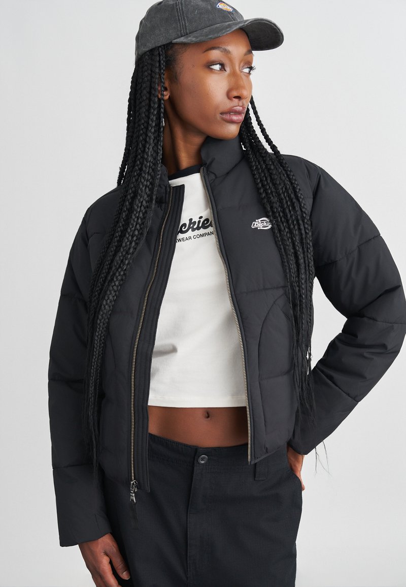 Giacca a vento nera con zip frontale, logo bianco, design cropped e polsini a costine. Indossata sopra una maglietta corta bianca e abbinata a pantaloni scuri.