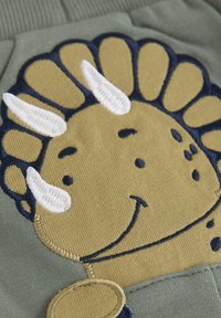 Groene stof met een beige geborduurd figuur met een glimlachend gezicht, witte hoorns en blauwe accenten, met een opmerkelijke textuur.