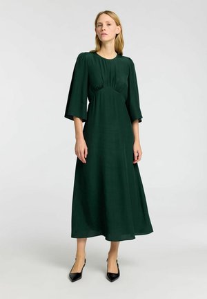 Robe midi verte à manches longues avec un buste ajusté et des manches évasées. Fabriquée en tissu lisse avec une texture subtile. Talons aiguilles noirs.