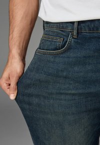 Tamsiai mėlynos džinsinės kelnės su klasikinio kirpimo, matomos priekinės kišenės ir detalės, tokios kaip siuvinėjimas bei audinio tekstūra. Rankos sukibusios už juosmens.