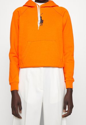 Personne portant un sweat à capuche orange vif avec un logo joueur de polo brodé noir et des cordons blancs, associé à un pantalon blanc taille haute.