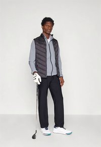 Colete acolchoado preto por cima de uma camisa cinza clara com fecho éclair, combinado com calças escuras e sapatos de golfe brancos. Segurando um taco de golfe, usando uma luva branca.