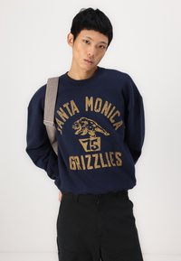 Sweatshirt azul-marinho com texto gráfico dourado "SANTA MONICA GRIZZLIES" e logótipo de urso; corte descontraído com decote redondo e punhos canelados.