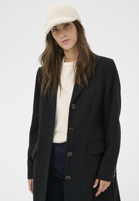 Black wool coat with a single-breasted design, featuring large black buttons, and a tailored fit. Paired with a light-colored shirt and a cream cap.