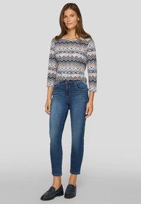 Frau steht und trägt ein langärmliges Oberteil mit Zickzackmuster, eine blaue Slim-Fit-Jeans und schwarze Slipper vor einem grauen Hintergrund.
