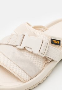 Beige sandaali, jossa on teksturoitu verkkomateriaali, säädettävä hihna muovisoljella ja keveä kumipohja. Brändin tagi näkyvissä.