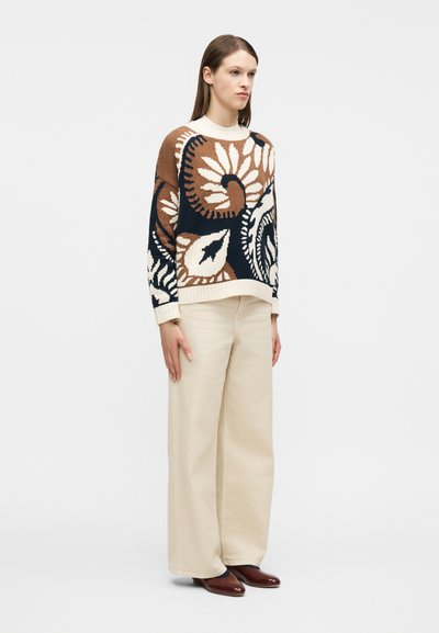 Γυναίκα που στέκεται φορώντας ένα πουλόβερ με καφέ, μαύρο και μπεζ λουλουδάτο μοτίβο, φαρδιά μπεζ παντελόνια και καφέ παπούτσια, με φόντο απλό λευκό.