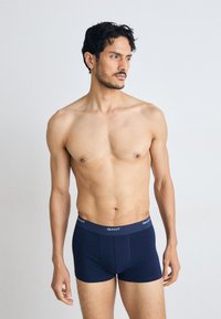 Μυώδης άνδρας με σκούρα μαλλιά που φοράει σκούρα μπλε μπόξερ GANT, στέκεται μπροστά σε ένα απλό ανοιχτό γκρι φόντο, κοιτάζοντας προς τα δεξιά του.