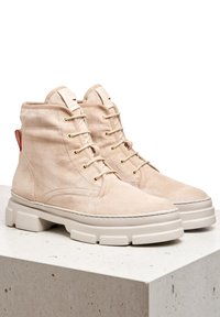 Botas de tobillo de ante beige con un acabado texturizado, puntera redonda, suela blanca gruesa y ojales en tono dorado, con un acento rojo en el talón.