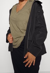 Persona vestida con una camiseta verde oliva de cuello en V debajo de una chaqueta negra parcialmente cerrada, acompañada de una falda negra.