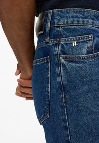 Vaqueros de denim en azul oscuro, con un diseño clásico de cinco bolsillos, detalles en acero inoxidable y un parche de cuero con el logo en la cintura.