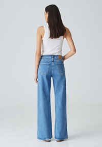 Jeans blu in denim a gamba larga con vita alta, dotati di una patch in pelle sul retro e due tasche posteriori. Indossati con un top bianco senza maniche.