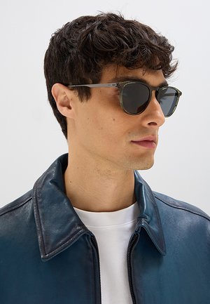 Uomo con occhiali da sole scuri, una giacca di pelle blu navy e una camicia bianca, mostrato in primo piano di profilo.